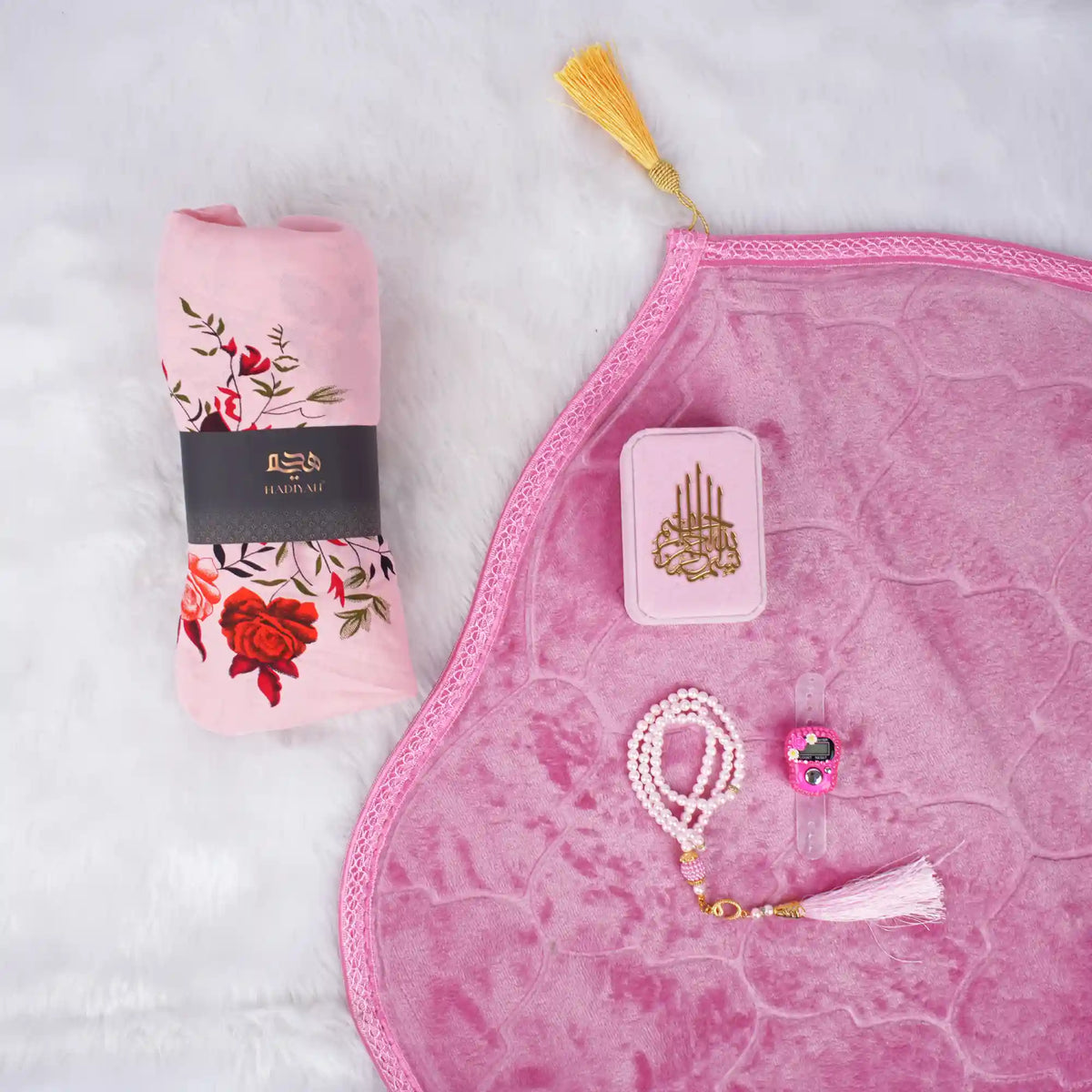 A soft pink Women's Namaz Essentials Hamper flat lay, featuring a rolled pink floral prayer dress, a pink velvet prayer mat, a pink mini velvet Quran box, white and pink tasbeeh beads, and a digital counter.