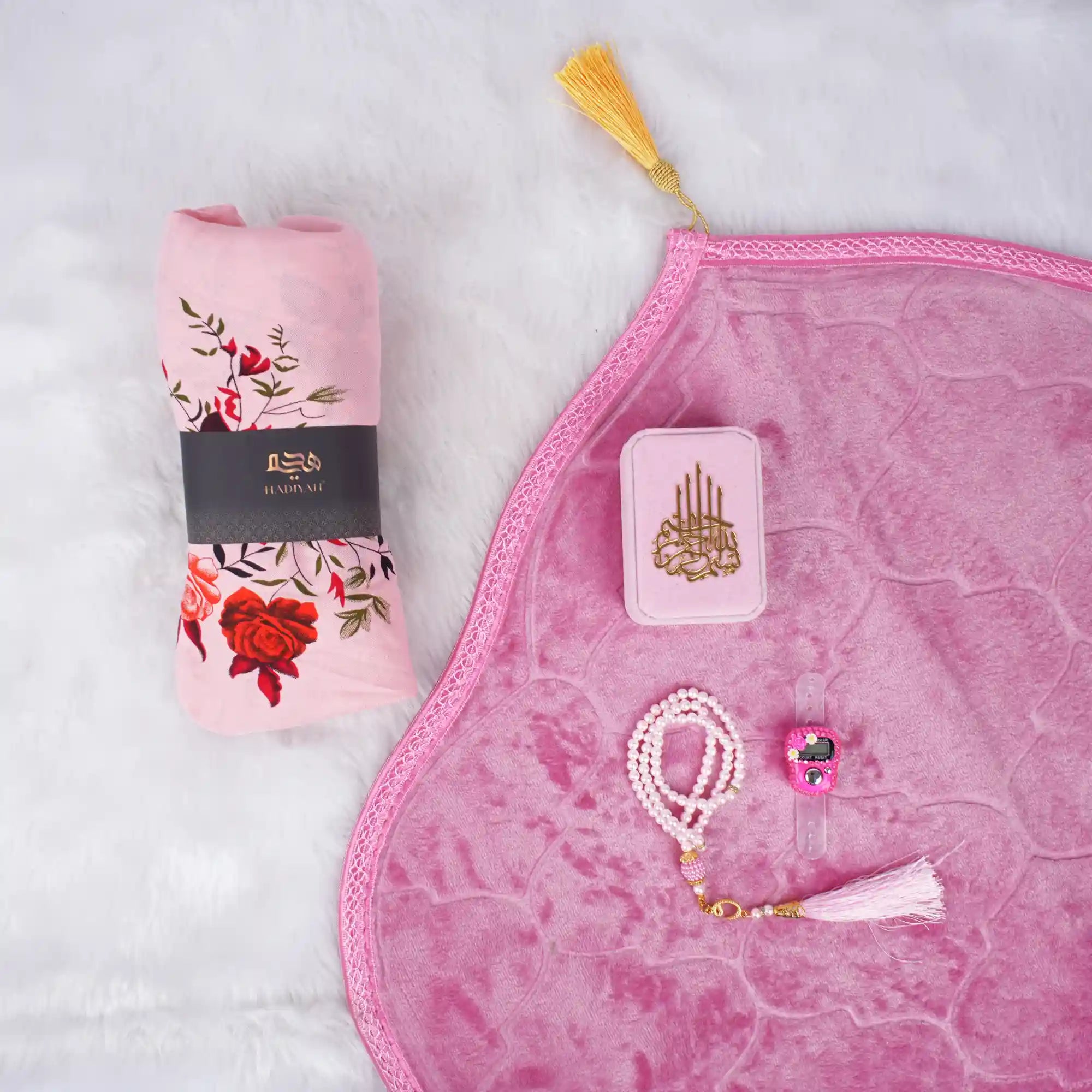 A soft pink Women's Namaz Essentials Hamper flat lay, featuring a rolled pink floral prayer dress, a pink velvet prayer mat, a pink mini velvet Quran box, white and pink tasbeeh beads, and a digital counter. Main image
