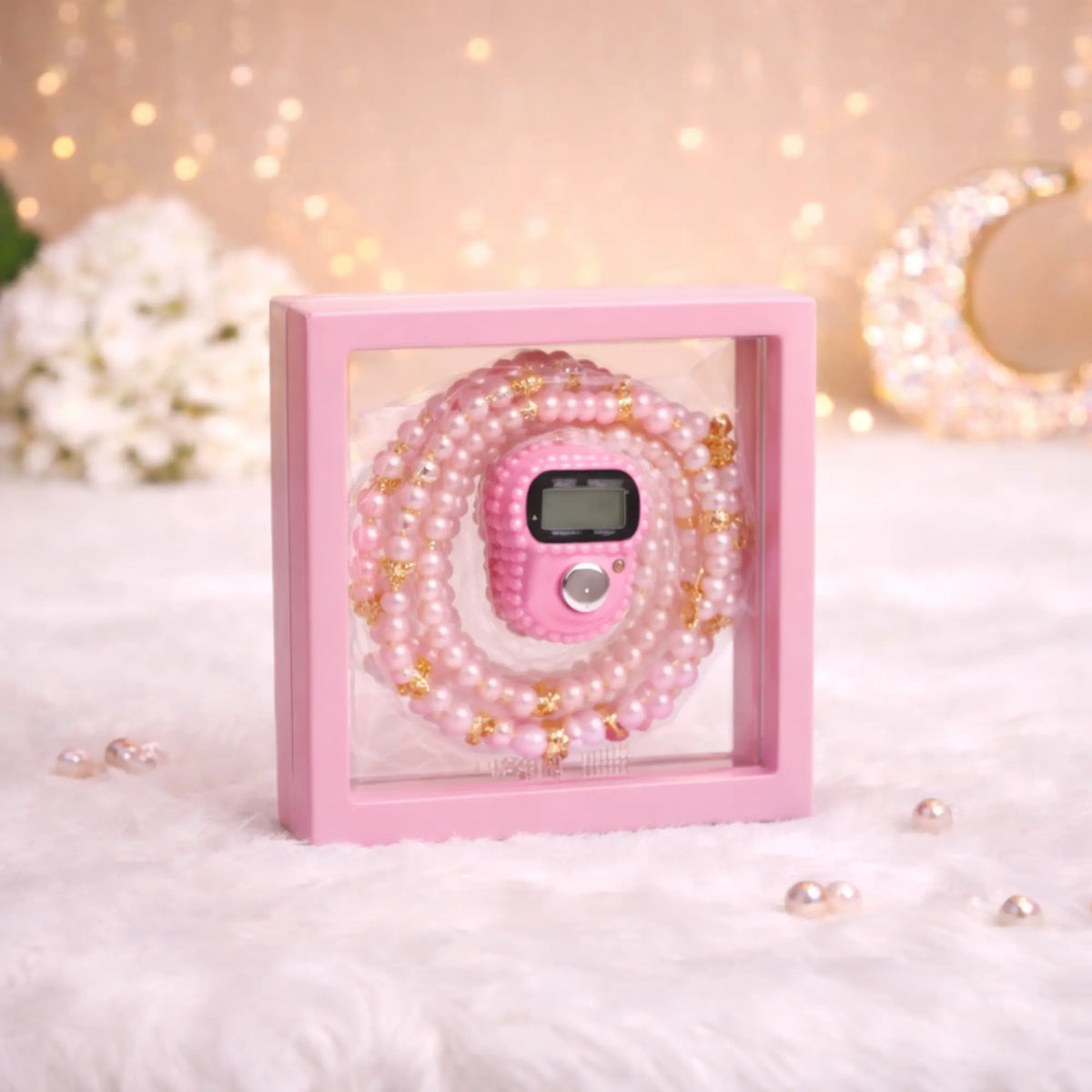Close-up of a pink digital tally counter and pink pearl tasbeeh beads beautifully presented inside a clear, pink-framed transparent display box. The box sits on a soft, fluffy white surface with a bokeh background of warm fairy lights and a crescent moon decoration.