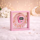 Close-up of a pink digital tally counter and pink pearl tasbeeh beads beautifully presented inside a clear, pink-framed transparent display box. The box sits on a soft, fluffy white surface with a bokeh background of warm fairy lights and a crescent moon decoration.