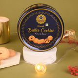 A close-up of a blue Kanha Danish Butter Cookies tin displayed upright on a white pedestal, surrounded by warm fairy lights and loose cookies against a green background.