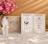 A styled wedding keepsake set featuring a cream-colored textured box with silver corner embellishments and a custom heart design, a matching decorated card with rhinestone corner accents, a white feather pen, and a cream tassel, arranged on a white faux fur surface with pink flowers in the background.