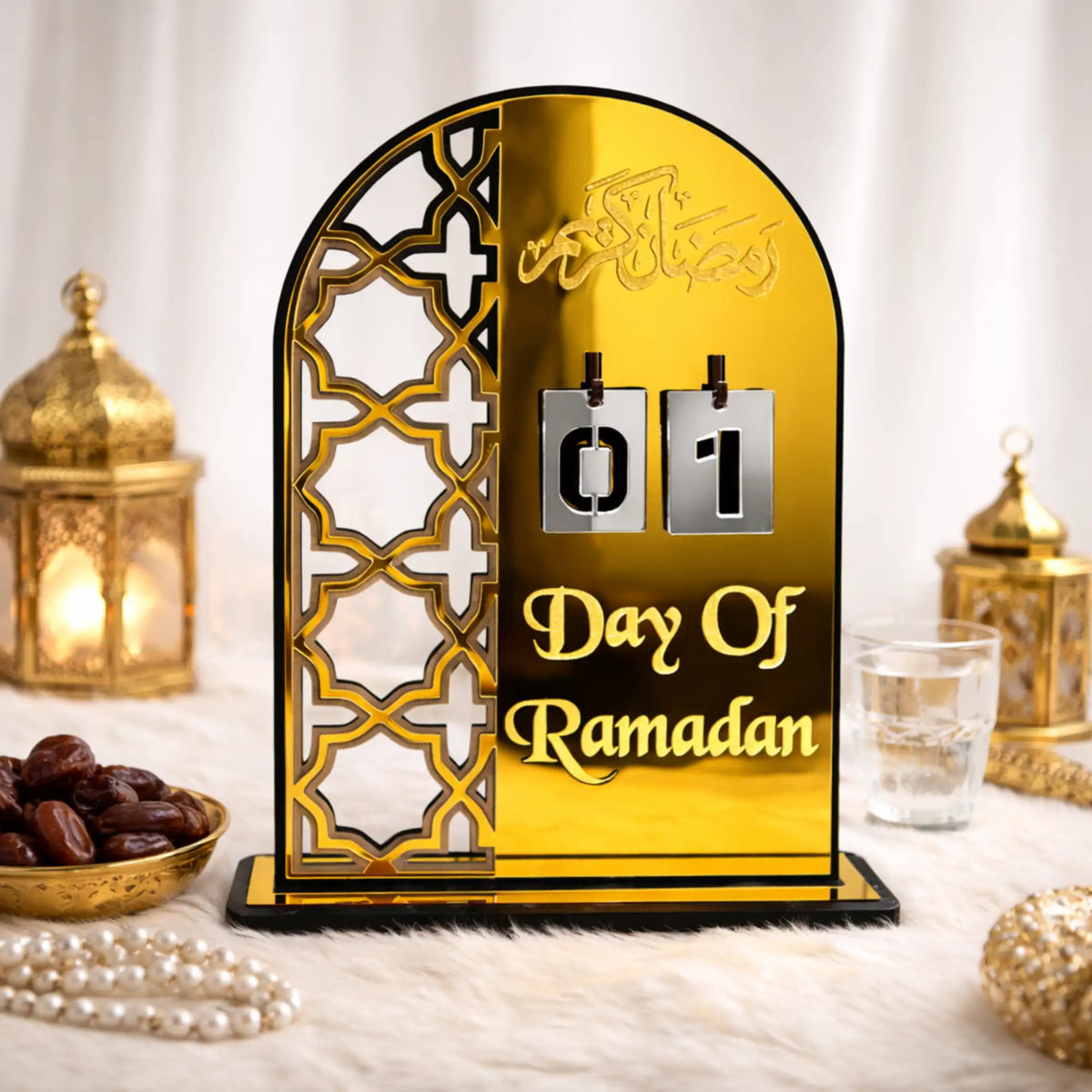 An arched gold acrylic Ramadan countdown calendar displaying the number 01 on hanging silver tiles above the text "Day Of Ramadan." The calendar features a cut-out geometric Islamic pattern on the left side and is displayed on a soft white surface alongside a bowl of dates, prayer beads, and gold lanterns.