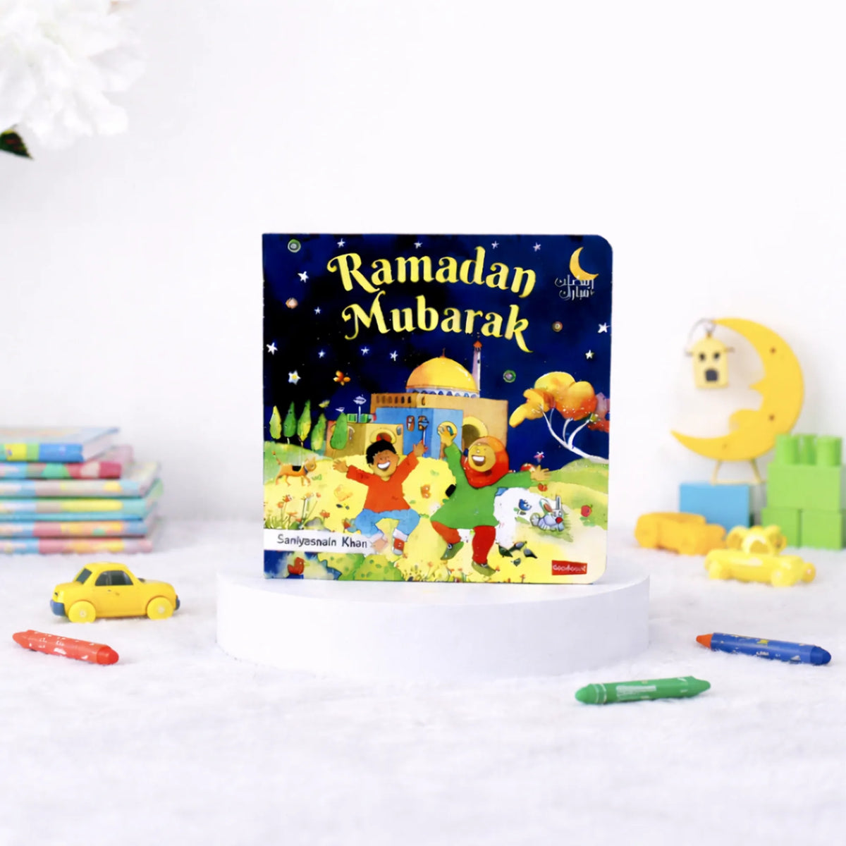 A children's board book titled "Ramadan Mubarak" by Saniyasnain Khan, resting on a white display stand. The colorful cover features children playing near a mosque under a starry night sky. The book is surrounded by scattered crayons, a small yellow toy car, and faintly blurred wooden toys in the background.