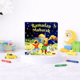 A children's board book titled "Ramadan Mubarak" by Saniyasnain Khan, resting on a white display stand. The colorful cover features children playing near a mosque under a starry night sky. The book is surrounded by scattered crayons, a small yellow toy car, and faintly blurred wooden toys in the background.