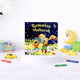 A children's board book titled "Ramadan Mubarak" by Saniyasnain Khan, resting on a white display stand. The colorful cover features children playing near a mosque under a starry night sky. The book is surrounded by scattered crayons, a small yellow toy car, and faintly blurred wooden toys in the background.