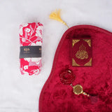 Red Sajdah Serenity Hamper displayed on a white fluffy surface, including a red velvet janamaz, a mini red velvet Quran, a red crystal tasbeeh, and a rolled-up red and white floral prayer makhana.