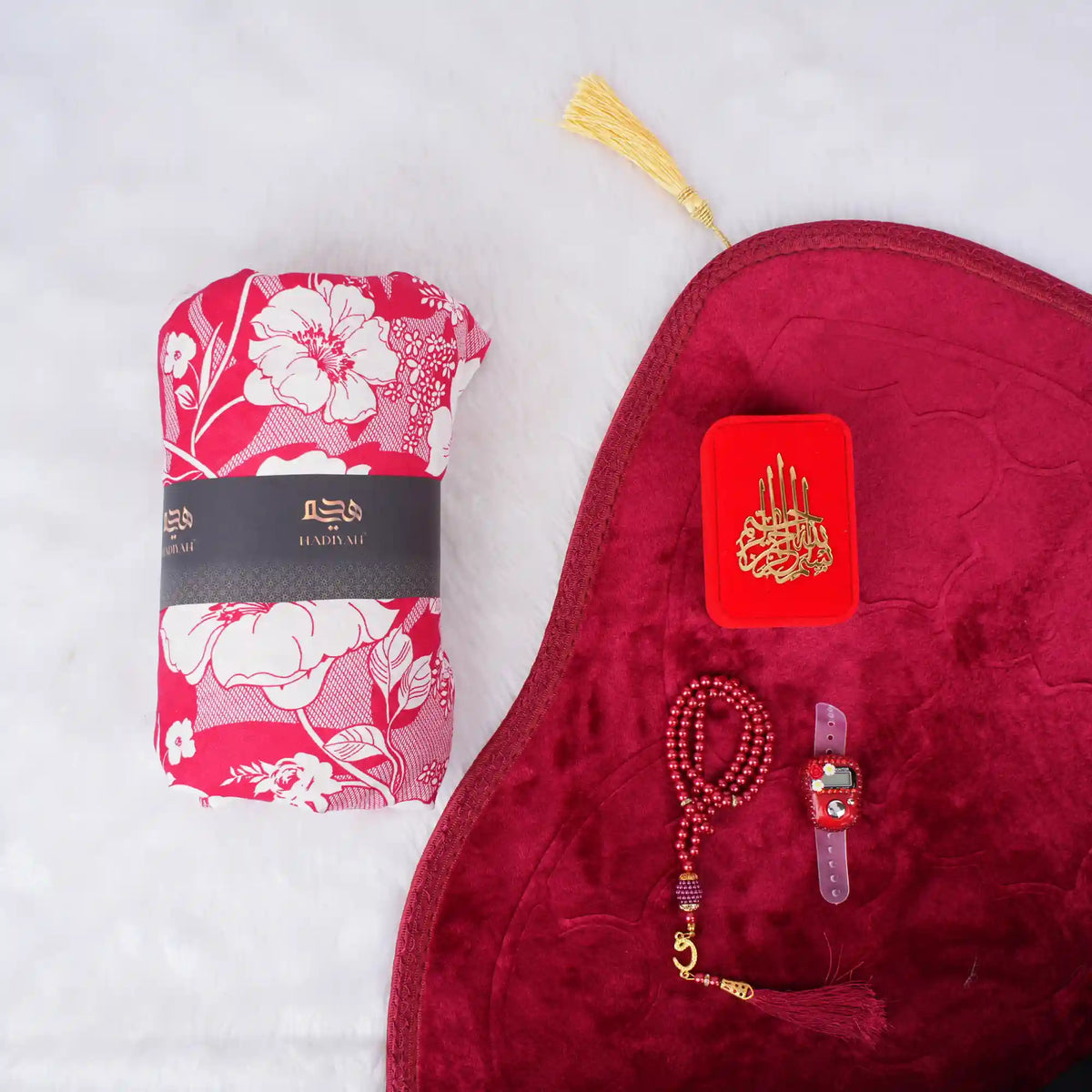 A red-themed Women's Namaz Essentials Hamper shown from above, including a rolled floral prayer dress, a red velvet prayer mat, a red mini velvet Quran box, matching red tasbeeh beads, and a digital counter.