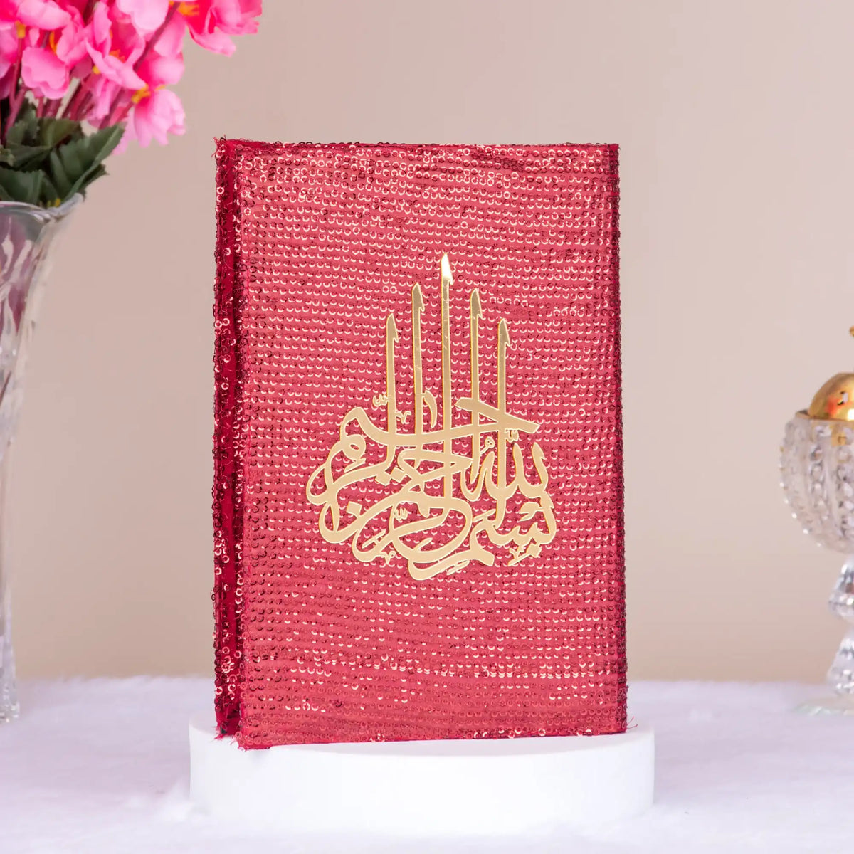 Red decorative book with gold Arabic calligraphy on a white surface with pink flowers in the background