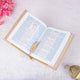 Open book with a gold bookmark on a light pink surface with pink flowers and a gold decorative item.