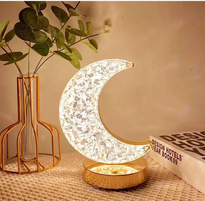 Decorative moon-shaped lamp on a table with a vase and book in the background Main image