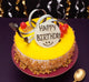 Round Birthday Butterscotch Cake topped with chocolate ribbons, cherries, and a 'Happy Birthday' plaque, ideal for festive celebrations.