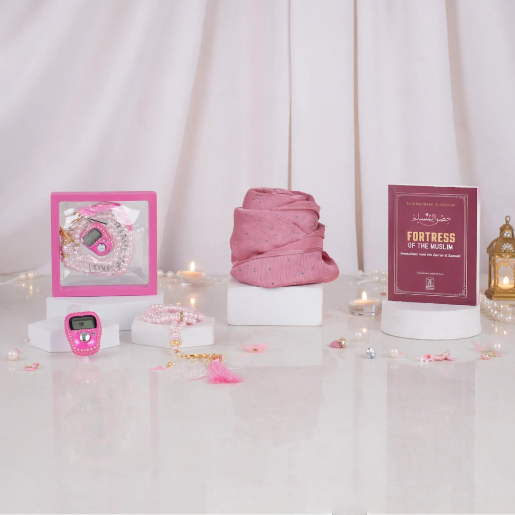 Front display of a pink Graceful Hijab Set women's hamper resting on white display blocks over a glossy surface scattered with pearls and pink petals. The set features a rolled pink hijab, a dark red "Fortress of the Muslim" dua book, a framed pink digital tasbeeh, a pink pearl tasbeeh with a tassel, and a matching tally counter, set against white curtains, fairy lights, and a golden lantern. Main image