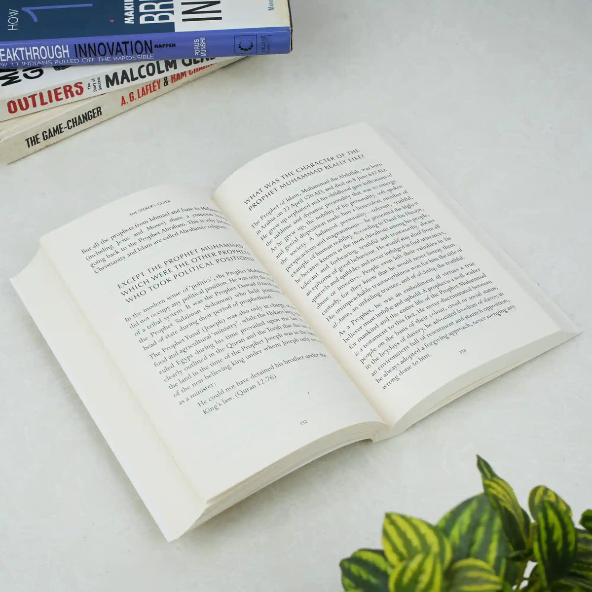 Open book on a white surface with other books stacked in the background