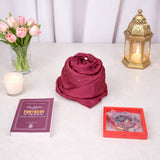 Top-down view of a red Graceful Hijab Set women's hamper featuring a dark red diamante hijab rolled into a rose shape, a purple "Fortress of the Muslim" dua book, and a red digital tasbeeh in a display frame. The items are arranged on a white marble surface next to a vase of pink tulips, a lit candle, and a golden decorative lantern.