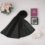 Flat lay view of a black Graceful Hijab Set featuring a beautifully draped black hijab with scattered diamantes, a purple "Fortress of the Muslim" dua book, and a black digital tasbeeh in a display box. The set is arranged on a white surface alongside a vase of pink roses, a lit candle in a glass jar, and a string of white pearls.