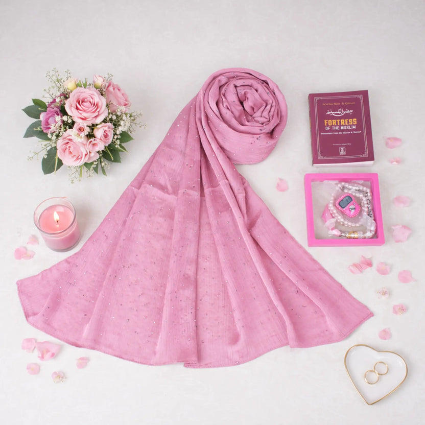 Flat lay view of a pink Graceful Hijab Set featuring a draped pink diamante hijab, a dark red "Fortress of the Muslim" dua book, and a pink digital tasbeeh inside a display frame. The items are spread across a white surface adorned with scattered pink petals, a bouquet of pink roses, a lit pink candle, and a heart-shaped ring dish. Main image