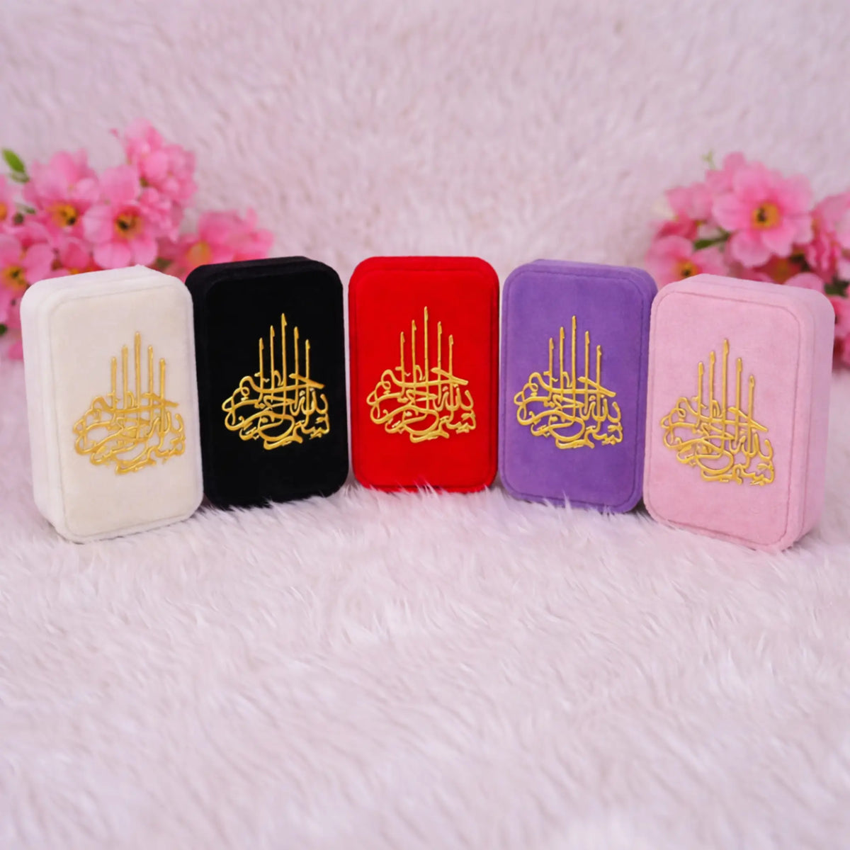 Five mini velvet Quran boxes lined up in a row in colors off-white, black, red, purple, and pink. Each box is decorated with gold Arabic calligraphy.