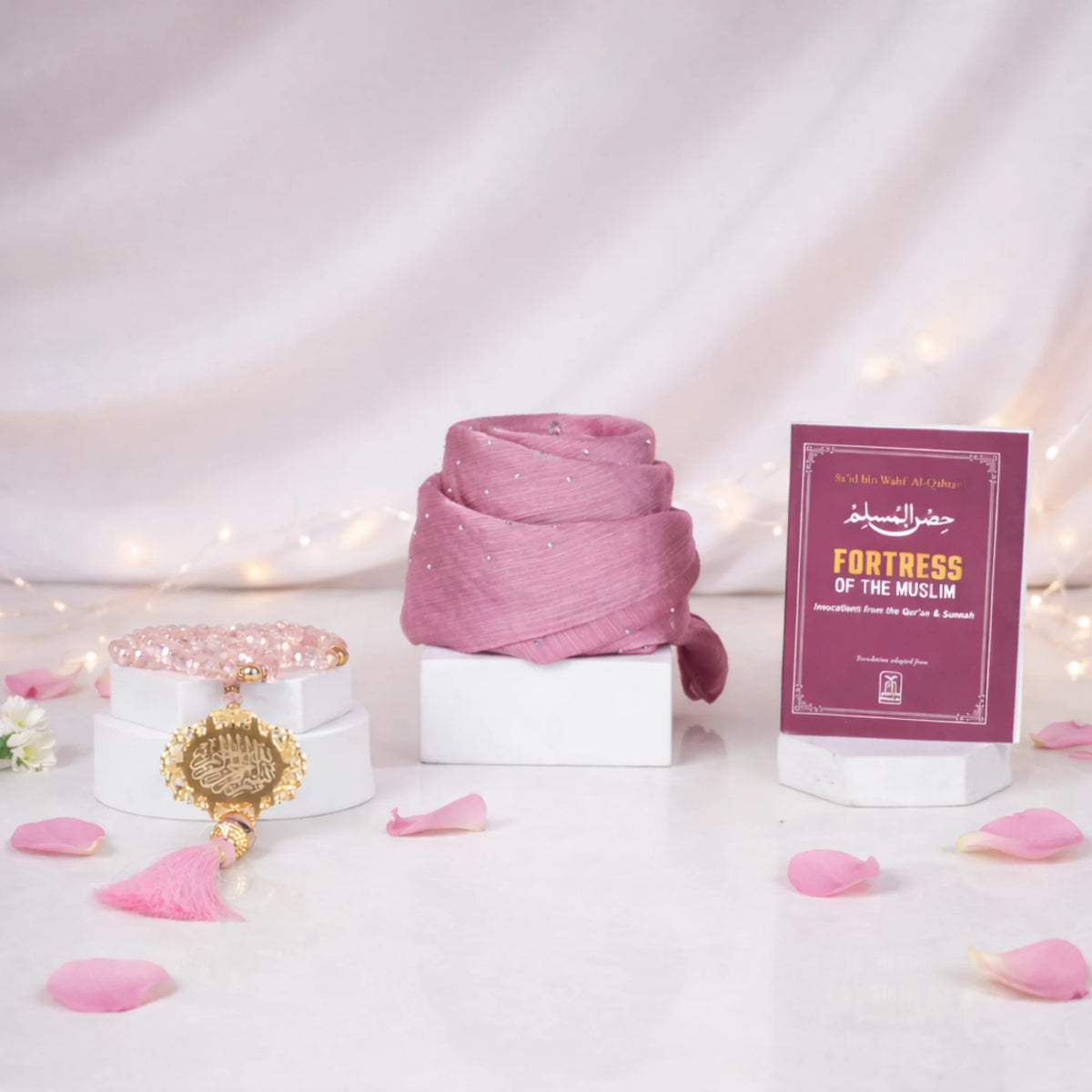 Front display of the Amara Elegance Women's Hamper featuring a neatly rolled pink rhinestone hijab, a pink crystal tasbeeh with a gold pendant and tassel, and a maroon "Fortress of the Muslim" Dua Book. The items are beautifully arranged on white display blocks against a soft white draped background with string lights and scattered pink rose petals.