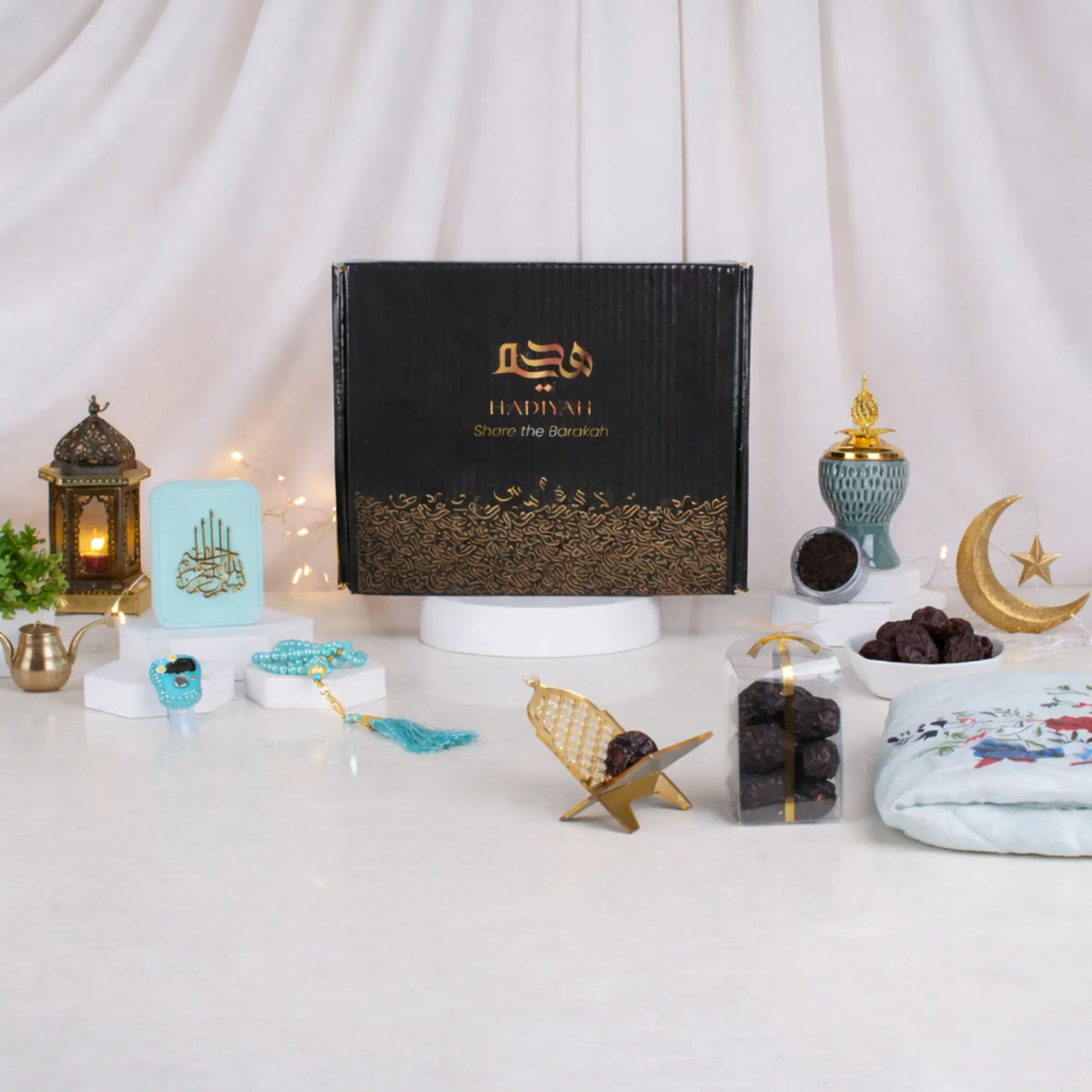 A front-facing display of the Velvet Serenity Women's Hamper in a blue theme. A black gift box with gold calligraphy is surrounded by a light blue floral prayer makhna, a cyan velvet tasbeeh box, a jade crown bakhoor burner, Ajwa dates, and a gold acrylic date stand against a draped white background with soft string lights.