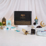 A front-facing display of the Velvet Serenity Women's Hamper in a blue theme. A black gift box with gold calligraphy is surrounded by a light blue floral prayer makhna, a cyan velvet tasbeeh box, a jade crown bakhoor burner, Ajwa dates, and a gold acrylic date stand against a draped white background with soft string lights.