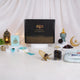 A front-facing display of the Velvet Serenity Women's Hamper in a blue theme. A black gift box with gold calligraphy is surrounded by a light blue floral prayer makhna, a cyan velvet tasbeeh box, a jade crown bakhoor burner, Ajwa dates, and a gold acrylic date stand against a draped white background with soft string lights.