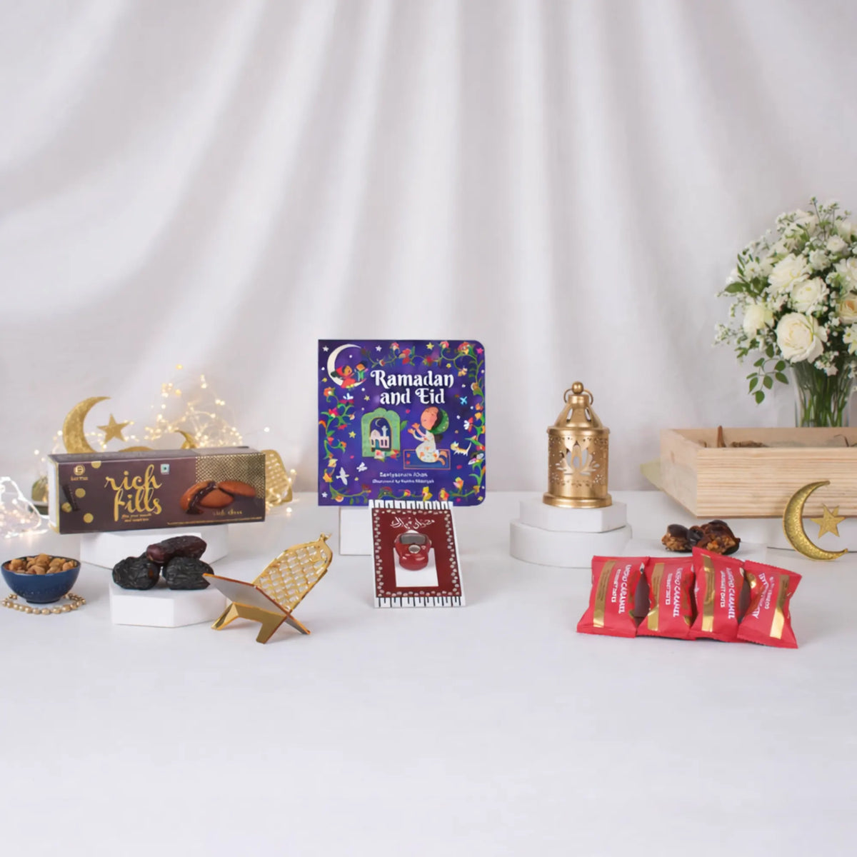 The individual items of the Festive Noor Kids Hamper spread out beautifully against a white draped background. The festive arrangement features a 'Ramadan and Eid' children's book, a box of Rich Fills cookies, a digital tasbeeh mounted on a themed card, a golden mini lantern, a golden acrylic date stand, four red-wrapped Loyka date chocolates, fresh dates on a pedestal, and a small blue bowl of nuts. The scene is accented with warm fairy lights, a vase of white roses, and a golden crescent moon decoration.