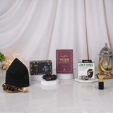 A front-view display of the Gentlemen's Essence Tray Hamper, showcasing its components: a black kufi (prayer cap), Ajwa dates, a maroon 'Fortress of the Muslim' book, a Zikr Ring in its box, and a small attar perfume bottle, arranged on geometric stands with a decorative lantern, against a curtained backdrop.