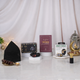 A front-view display of the Gentlemen's Essence Tray Hamper, showcasing its components: a black kufi (prayer cap), Ajwa dates, a maroon 'Fortress of the Muslim' book, a Zikr Ring in its box, and a small attar perfume bottle, arranged on geometric stands with a decorative lantern, against a curtained backdrop.