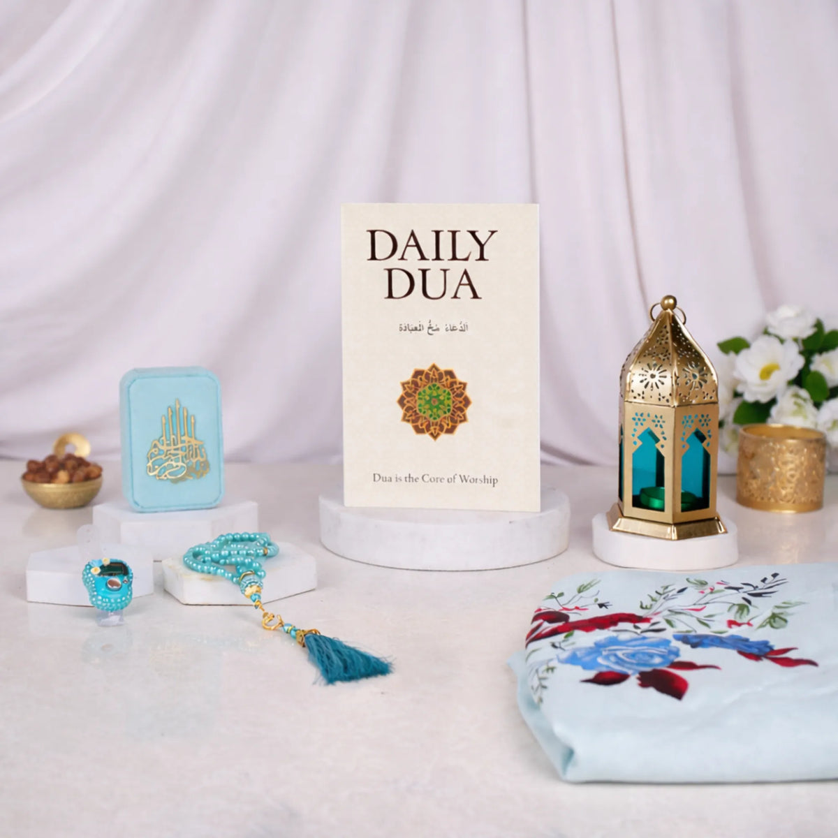 An elegant, styled display of the Grace & Glow Women's Hamper contents against a softly draped white fabric background. A Daily Dua book stands upright in the center on a small marble pedestal. To its left is a light blue velvet box, a blue digital ring counter, and a blue beaded tasbeeh with a tassel. To the right is a gold and blue metal lantern. A portion of the light blue floral prayer makhna rests in the right foreground.