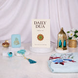 An elegant, styled display of the Grace & Glow Women's Hamper contents against a softly draped white fabric background. A Daily Dua book stands upright in the center on a small marble pedestal. To its left is a light blue velvet box, a blue digital ring counter, and a blue beaded tasbeeh with a tassel. To the right is a gold and blue metal lantern. A portion of the light blue floral prayer makhna rests in the right foreground.