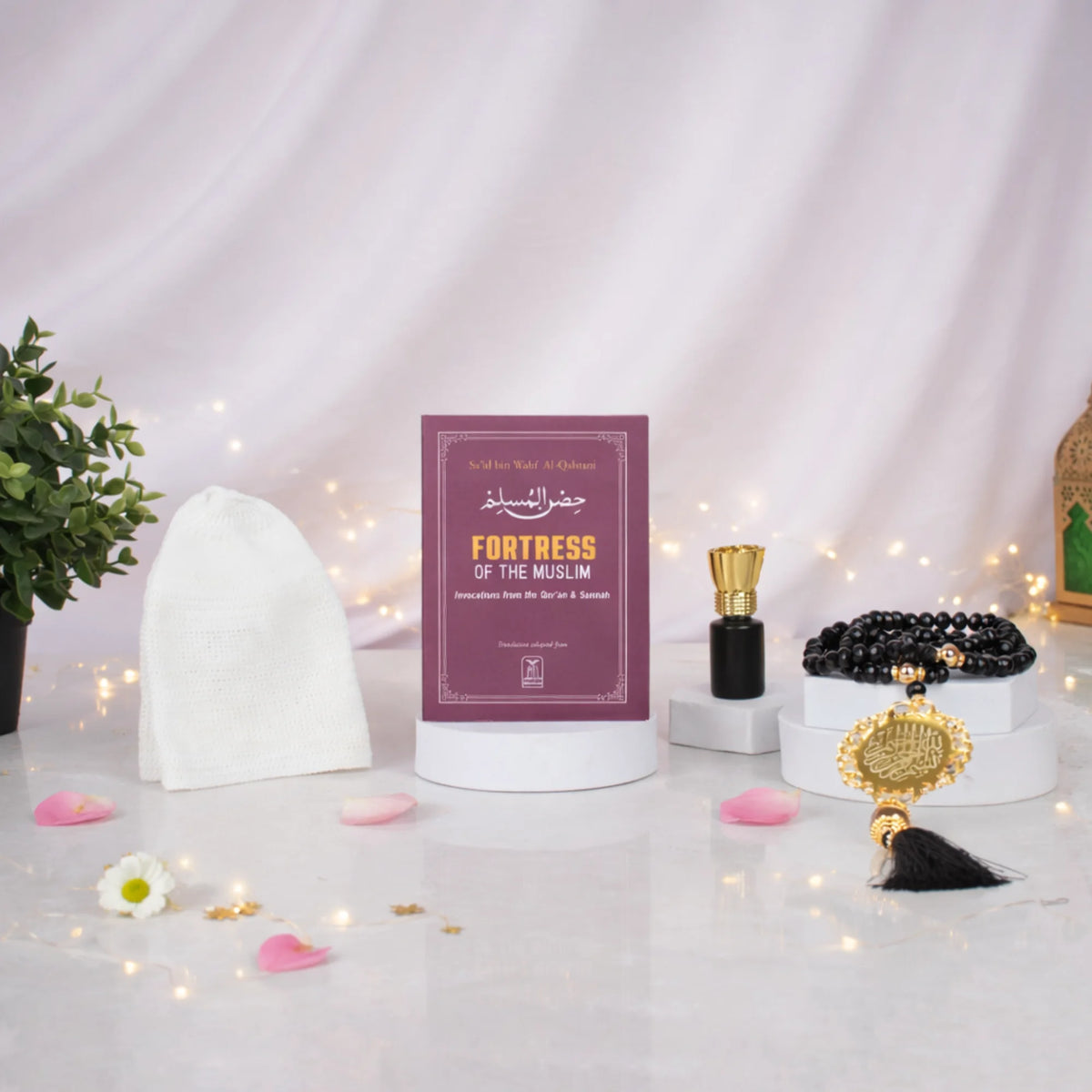 Front display of the Imaan Prestige Men's Hamper arranged on small white pedestals. A maroon Fortress of the Muslim dua book stands in the center, flanked by a white prayer cap, a bottle of attar, and black crystal prayer beads with a gold pendant. The setup is set against a draped white fabric background with warm fairy lights and scattered pink rose petals.