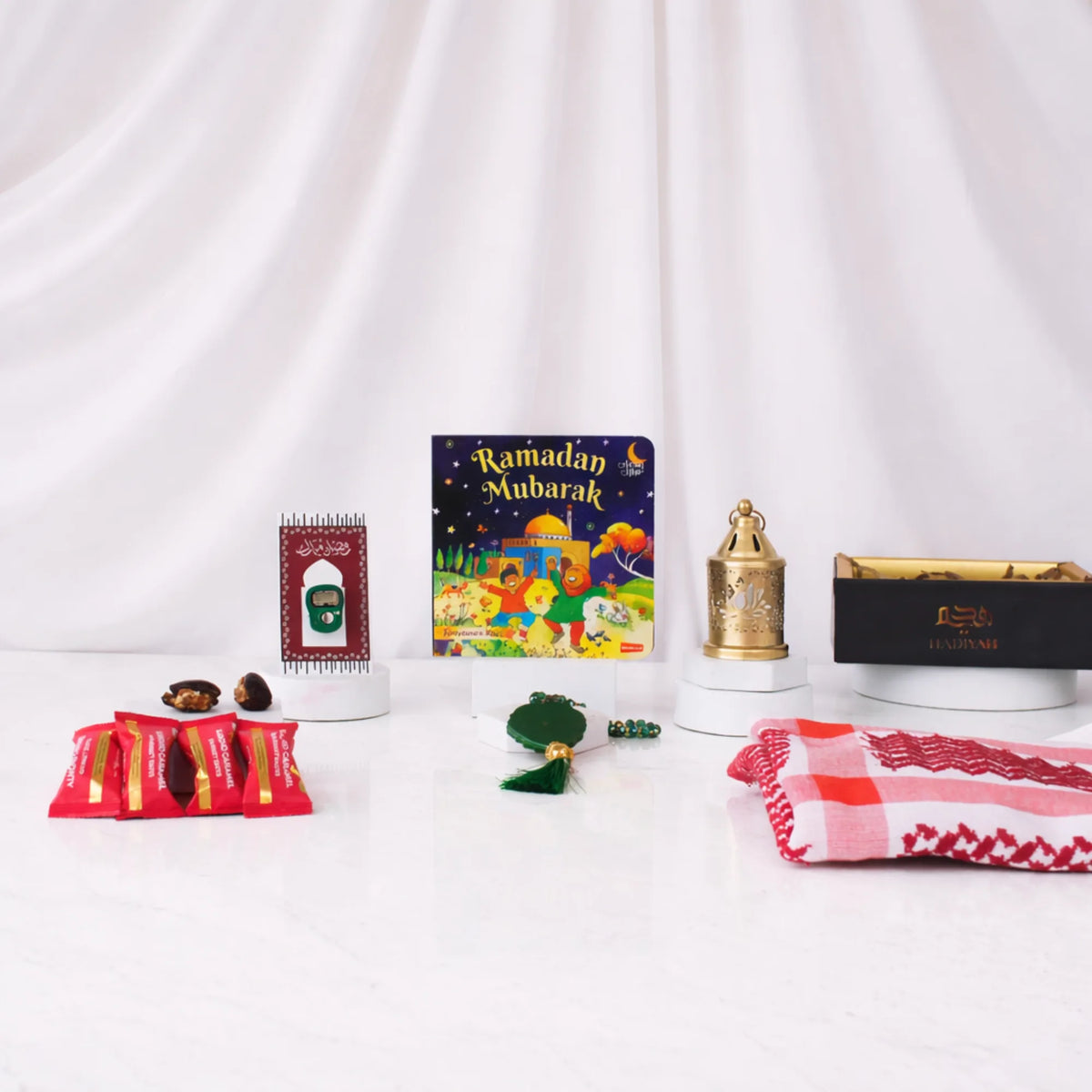 Front view of the Little Charm Tray Hamper items beautifully displayed against a white draped fabric background. The arrangement includes a "Ramadan Mubarak" children's board book in the center, a green digital tasbeeh mounted on a decorative card, four red-wrapped date chocolates, a green crystal 33-bead tasbeeh in the foreground, a mini golden lantern on a small pedestal, a folded red and white patterned prayer scarf, and a black and gold Hadiyah gift tray.