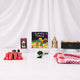 Front view of the Little Charm Tray Hamper items beautifully displayed against a white draped fabric background. The arrangement includes a "Ramadan Mubarak" children's board book in the center, a green digital tasbeeh mounted on a decorative card, four red-wrapped date chocolates, a green crystal 33-bead tasbeeh in the foreground, a mini golden lantern on a small pedestal, a folded red and white patterned prayer scarf, and a black and gold Hadiyah gift tray.