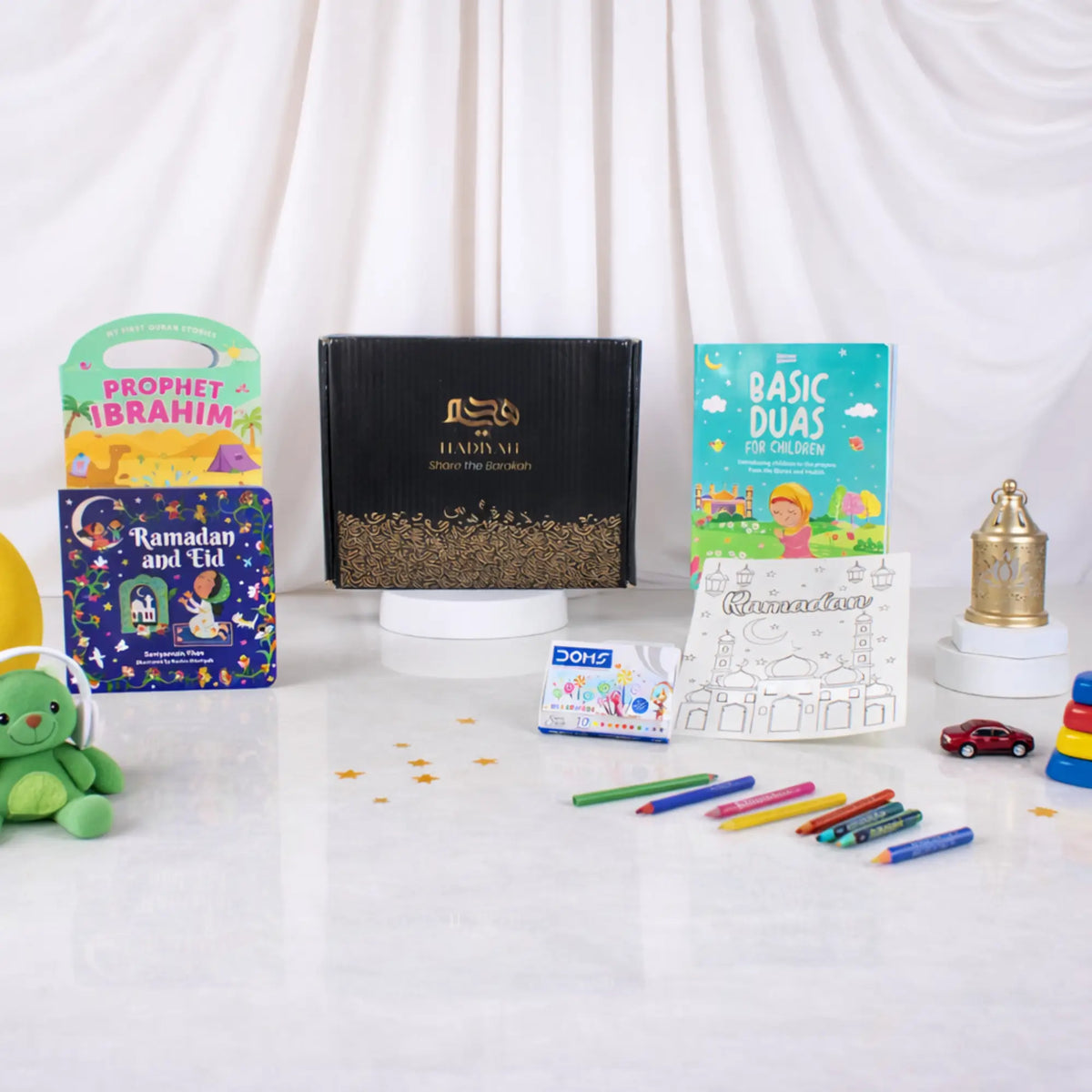 A front display of the Little Scholars of Faith Hamper contents spread on a white surface, featuring a black Hadiyah gift box, three children's Islamic books (Prophet Ibrahim, Ramadan and Eid, and Basic Duas), a Ramadan colouring sheet, a pack of crayons, scattered gold stars, and a mini golden lantern.