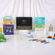 A front display of the Little Scholars of Faith Hamper contents spread on a white surface, featuring a black Hadiyah gift box, three children's Islamic books (Prophet Ibrahim, Ramadan and Eid, and Basic Duas), a Ramadan colouring sheet, a pack of crayons, scattered gold stars, and a mini golden lantern.