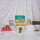 A front-facing display of a children's Ramadan gift set arranged on a white surface. The collection includes "The Story of Maryam" book, a white crocheted prayer cap, a green digital tasbeeh, red wrapped date chocolates, a green crystal tasbeeh with a large tassel, a decorative golden lantern, and a black gift box. The items are set against a softly draped white fabric illuminated by warm fairy lights.