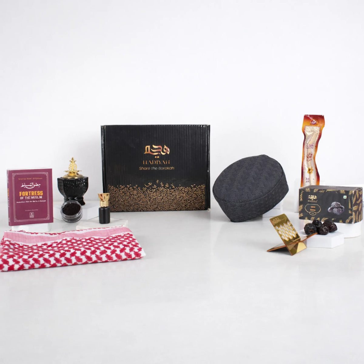 Front display of the Noble Ibadah Men's Hamper by Hadiyah, featuring a black gift box surrounded by a ghutra, Islamic book, bakhoor burner, prayer cap, miswak, and Ajwa dates.