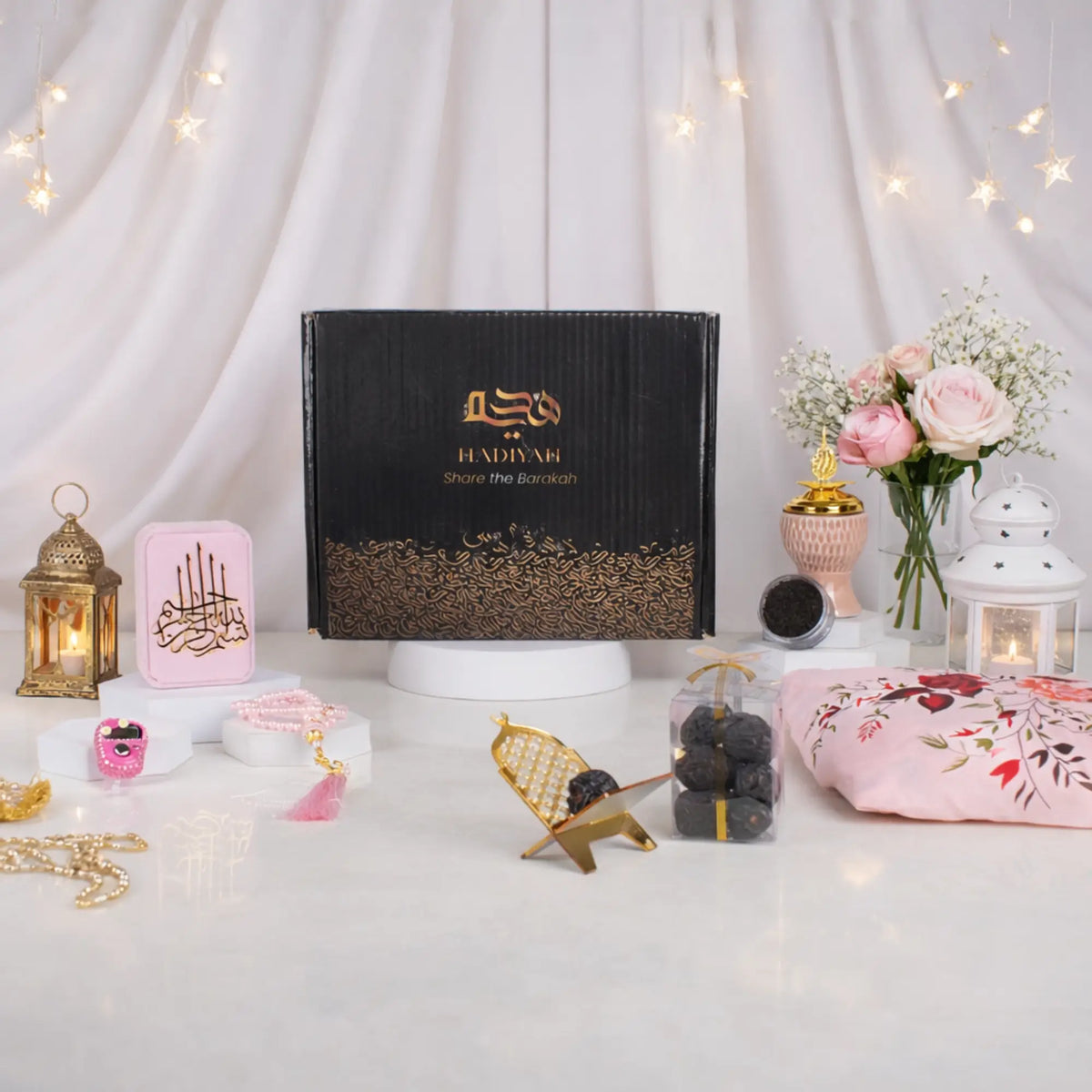 A front-facing display of the Velvet Serenity Women's Hamper in a pink theme. A black and gold gift box is surrounded by a pink floral prayer makhna, a pink velvet tasbeeh box with matching prayer beads, a blush pink crown bakhoor burner, Ajwa dates, and a gold acrylic date stand, arranged elegantly among decorative lanterns and flowers.