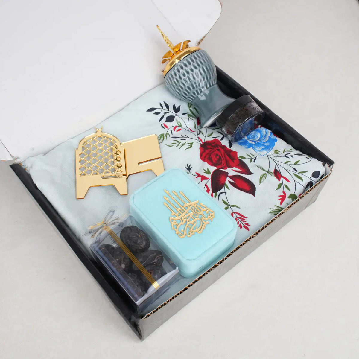 A top-down view of the open Velvet Serenity Women's Hamper box, showcasing the blue-themed contents neatly packed inside. A light blue floral prayer makhna forms the base, topped with a cyan velvet tasbeeh box, a gold acrylic date stand, a clear box of Ajwa dates, and a jade crown bakhoor burner.