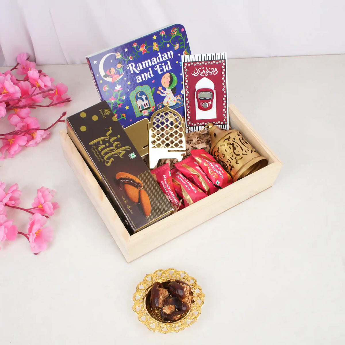 An angled top-down view of the Festive Noor Kids Hamper neatly arranged inside a light square wooden tray. Nestled on a bed of brown crinkle paper are a 'Ramadan and Eid' book, a box of Rich Fills cookies, a digital tasbeeh card, a golden mini lantern, an acrylic date stand, and red-wrapped date chocolates. A sprig of pink artificial cherry blossoms and a small golden decorative dish holding stuffed dates sit beside the tray on a white surface.