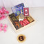 An angled top-down view of the Festive Noor Kids Hamper neatly arranged inside a light square wooden tray. Nestled on a bed of brown crinkle paper are a 'Ramadan and Eid' book, a box of Rich Fills cookies, a digital tasbeeh card, a golden mini lantern, an acrylic date stand, and red-wrapped date chocolates. A sprig of pink artificial cherry blossoms and a small golden decorative dish holding stuffed dates sit beside the tray on a white surface.