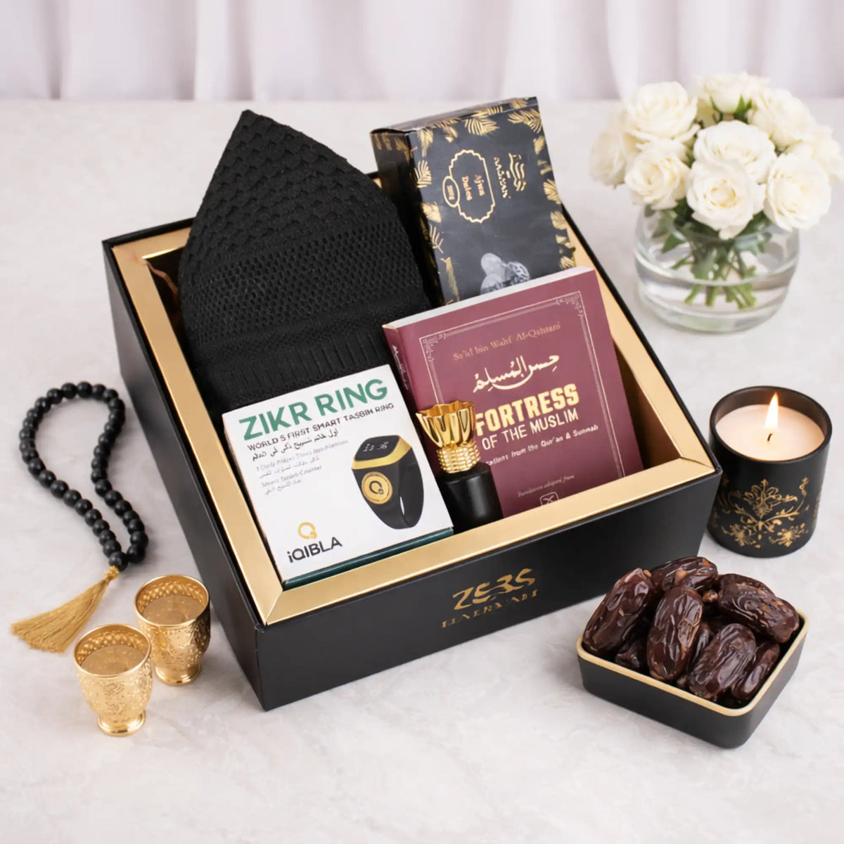 A top-down, slightly angled view of the packed Gentlemen's Essence Tray Hamper in its black presentation box with gold trim. The items visible are the black kufi, Ajwa dates, the 'Fortress of the Muslim' book, Zikr Ring box, and attar. It is surrounded by loose dates, two gold fincan cups, a lit candle, prayer beads, and white roses on a marble surface.