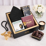 A top-down, slightly angled view of the packed Gentlemen's Essence Tray Hamper in its black presentation box with gold trim. The items visible are the black kufi, Ajwa dates, the 'Fortress of the Muslim' book, Zikr Ring box, and attar. It is surrounded by loose dates, two gold fincan cups, a lit candle, prayer beads, and white roses on a marble surface.