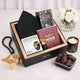 A top-down, slightly angled view of the packed Gentlemen's Essence Tray Hamper in its black presentation box with gold trim. The items visible are the black kufi, Ajwa dates, the 'Fortress of the Muslim' book, Zikr Ring box, and attar. It is surrounded by loose dates, two gold fincan cups, a lit candle, prayer beads, and white roses on a marble surface.