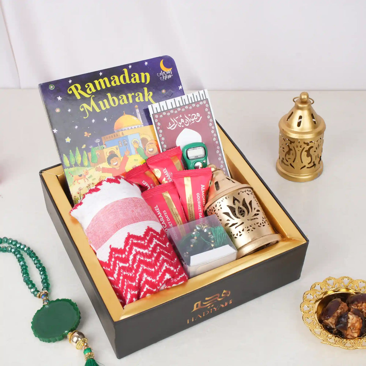 Angled top-down view of the Little Charm Tray Hamper beautifully packaged inside a black gift box with a gold inner border and the "HADIYAH" logo. The tray is filled with a "Ramadan Mubarak" board book, a folded red and white prayer scarf, red-wrapped date chocolates, a digital tasbeeh with its card, a mini golden lantern, and a small clear box containing a green crystal tasbeeh. An extra green tasbeeh, a golden lantern, and a decorative golden dish of dates are displayed on the white surface outside the bo