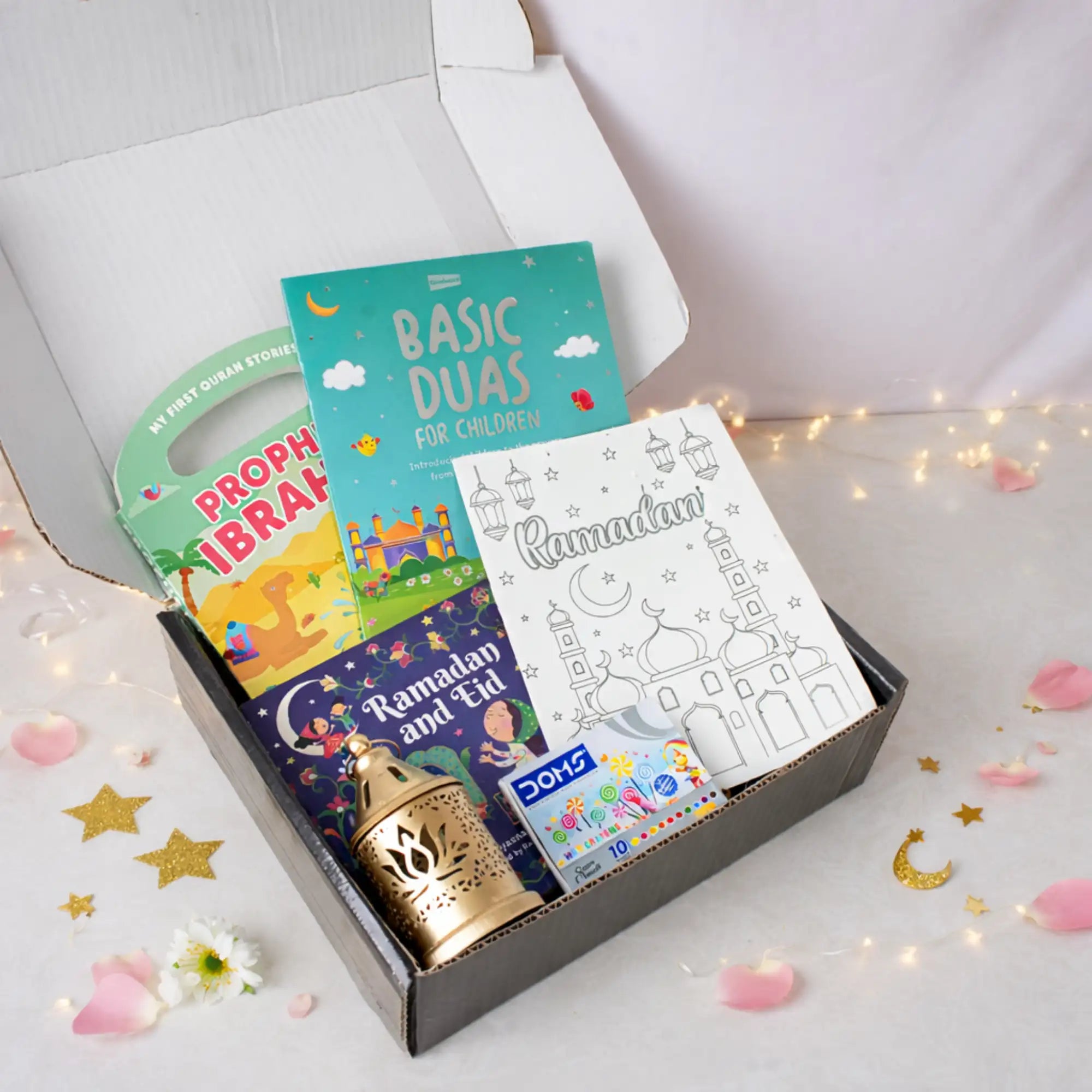 A top-down view of an open black gift box neatly packed with the Little Scholars of Faith Hamper items, including three children's books, a colouring sheet, crayons, and a mini golden lantern, surrounded by decorative fairy lights, pink petals, and gold star cutouts. Main image