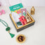 A top-down, angled view of a black and gold gift box neatly packed with children's Ramadan hamper items, including "The Story of Maryam" book, a white prayer cap, a digital tasbeeh, wrapped chocolates, and a mini golden lantern. Beside the box on the white surface lies a green crystal tasbeeh and a small, ornate golden dish filled with dates.