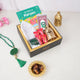 A top-down, angled view of a black and gold gift box neatly packed with children's Ramadan hamper items, including "The Story of Maryam" book, a white prayer cap, a digital tasbeeh, wrapped chocolates, and a mini golden lantern. Beside the box on the white surface lies a green crystal tasbeeh and a small, ornate golden dish filled with dates.