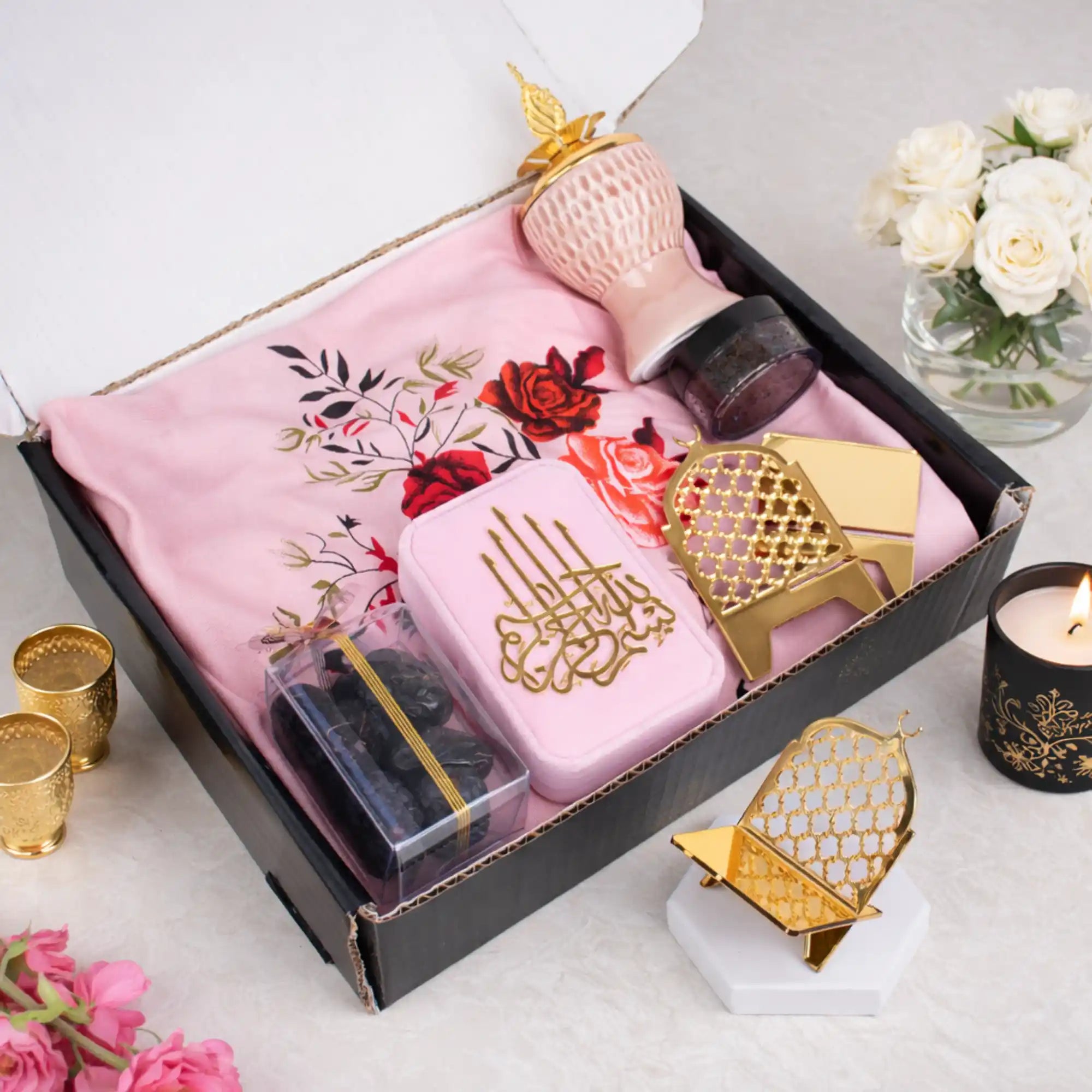 A top-down view of the open Velvet Serenity Women's Hamper box, revealing the pink-themed contents carefully arranged inside. A pink floral prayer makhna serves as the base layer, holding a pink velvet tasbeeh box with gold Arabic script, a gold acrylic date stand, a clear box of Ajwa dates, and a blush pink crown bakhoor burner. Main image